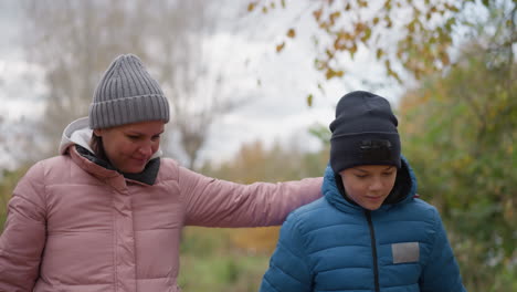 mother in pink jacket and son in blue jacket enjoy a walk together, she gently holds his arm; he playfully hits a tree branch with a stick, while she holds a leaf