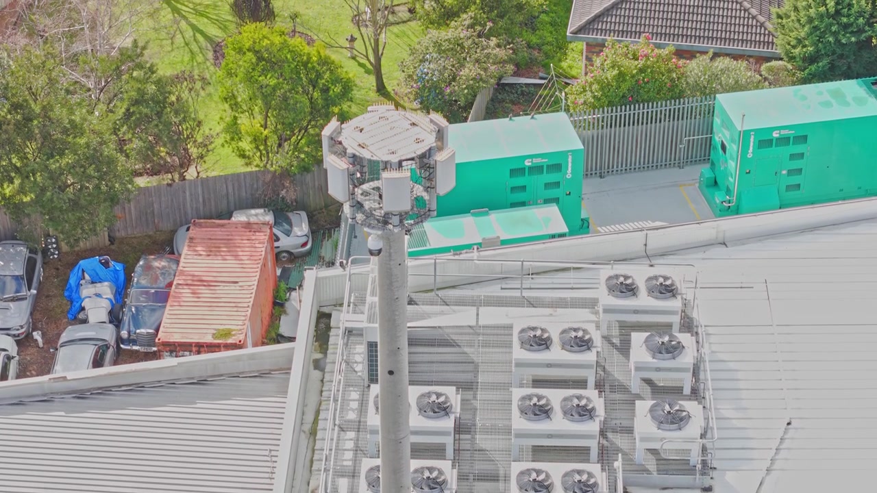 Drone video captures an overhead view of a modern data centre rooftop with industrial cooling units, backup generators, and parking area, in bright daylight and sharp focus.