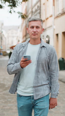 mature man using smartphone typing texting social media messages email while walking on city street