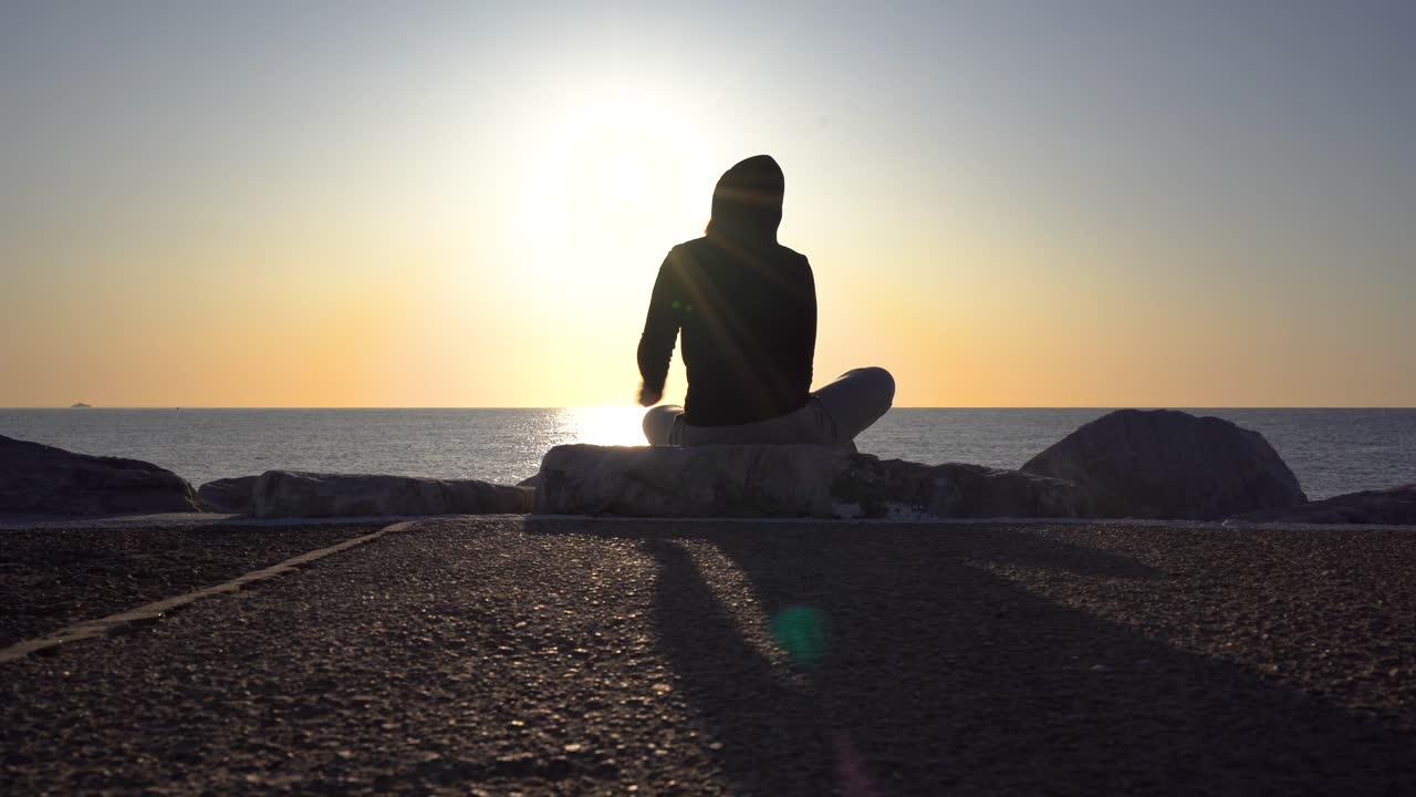 silueta joven con una sudadera con capucha en las rocas del muelle disfrutando del amanecer, ubicación puerto banús, marbella, málaga, españa, momento relajante, gimbal moviéndose a la derecha