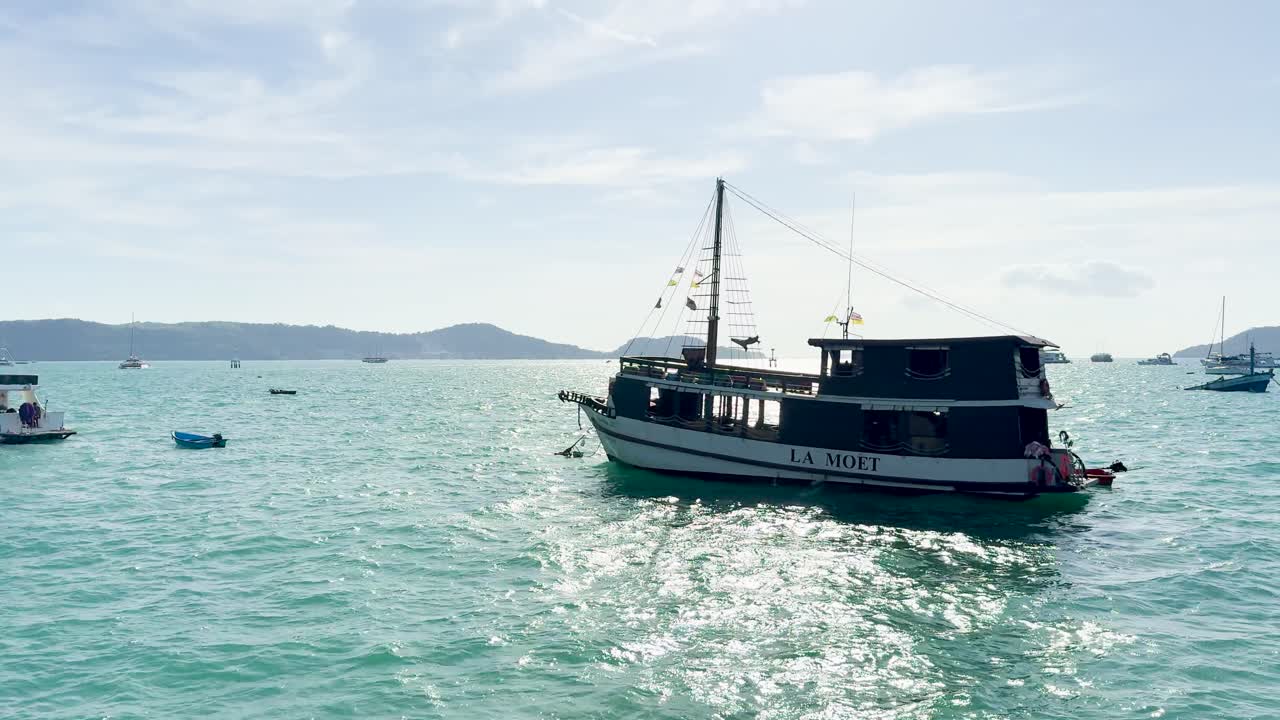 A boat glides through Chalong Bay, Phuket, under bright sunlight, surrounded by calm turquoise waters and distant islands