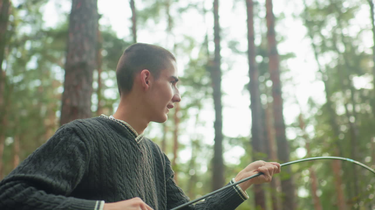 Natural light glow surrounds man in cable-knit sweater adjusting tent pole during forest camping as he prepares setup with assistance from partner, surrounded by tall trees and tranquil green scenery