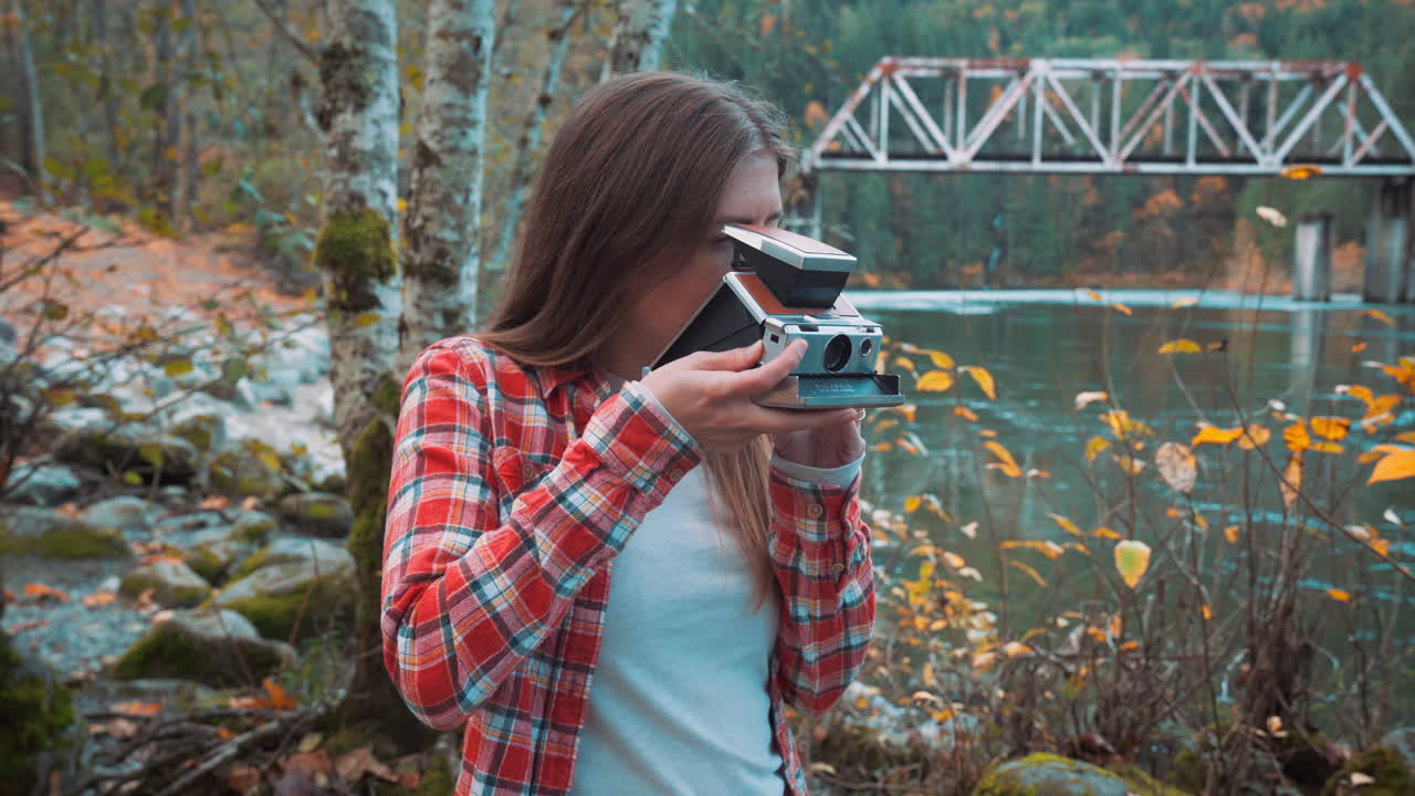 사진작가 여성이 가을 색상 중 강변을 따라 폴라로이드 사진을 찍습니다.