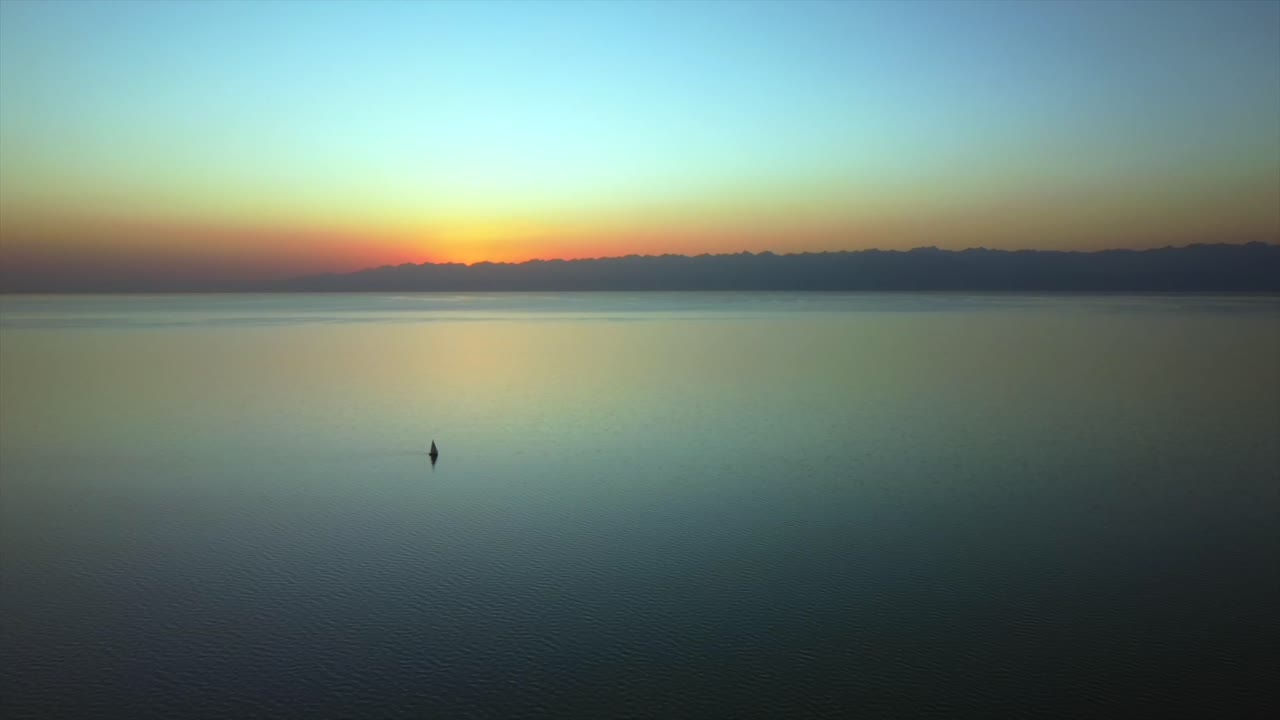 드론은 해질녘에 큰 호수 위를 날아다니며 멋진 일몰을 배경으로 탁 트인 전망에서 범선과 큰 산을 포착합니다.