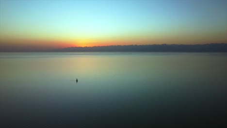 El-Dron-Vuela-Sobre-Un-Gran-Lago-Al-Atardecer,-Capturando-Un-Velero-Y-Grandes-Montañas-En-Una-Vista-Panorámica-Con-Los-Impresionantes-Colores-De-La-Puesta-De-Sol-En-El-Fondo