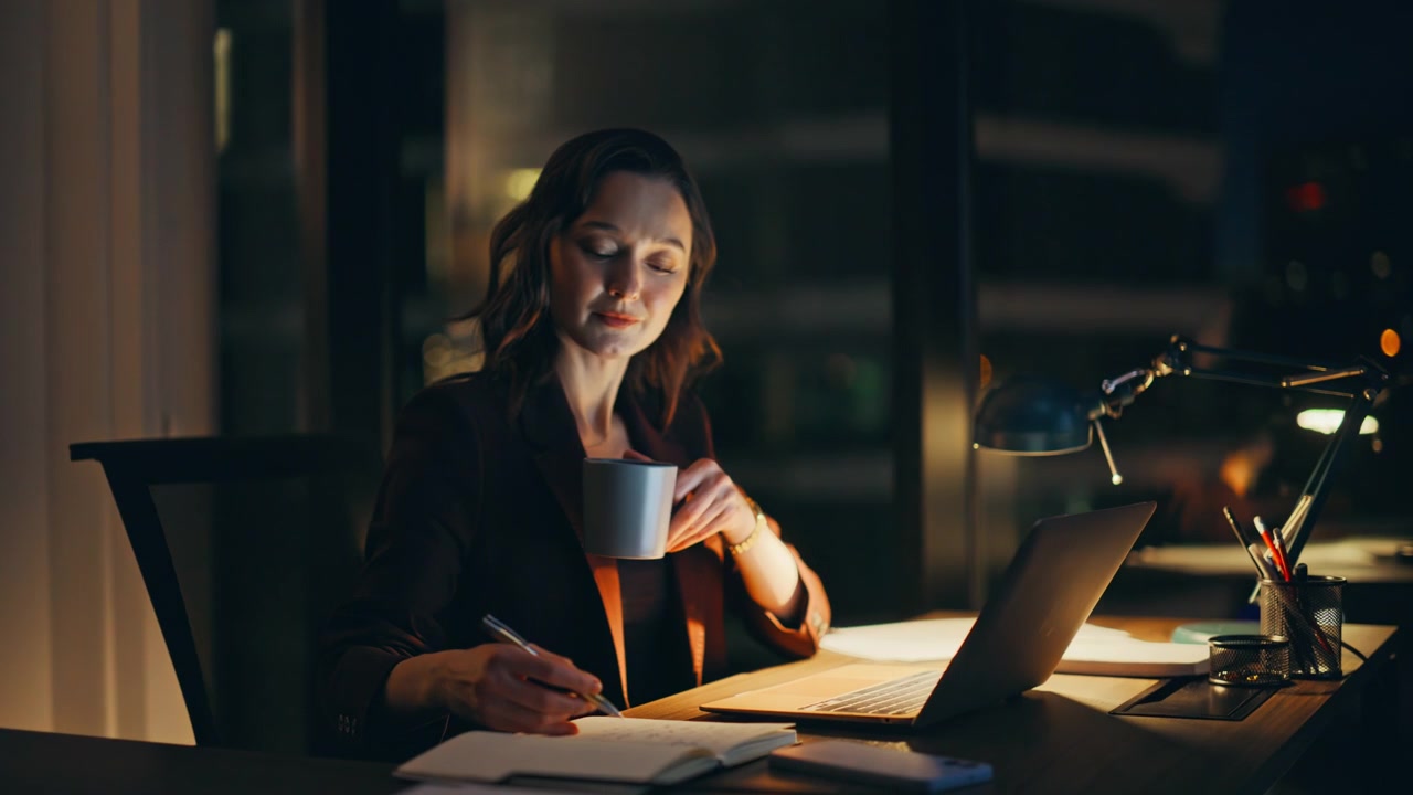 Manager felice che tiene una tazza di caffè, legge al computer e prende appunti in un ufficio buio