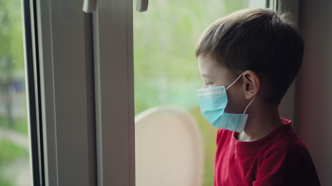 마스크를 입은 슬픈 어린 소년이 창문 근처에 앉아서 밖을 바라보고 있다.