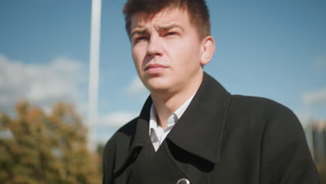 close-up of investor walking confidently outdoors checking time on wristwatch, wearing elegant black coat with metallic buttons, set against modern urban architecture, green trees, and clear blue sky