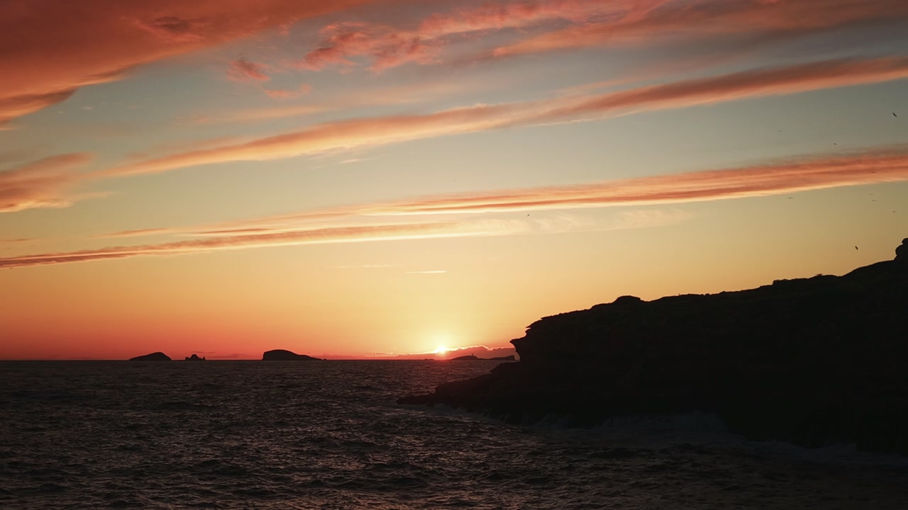 Drone panning and orbiting over Cala Comte revealing sunset over the sea with warm golden light on Ibiza coastline