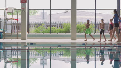 Female-Coach-Walks-Children-In-Swimming-Class-Along-Edge-Of-Indoor-Pool