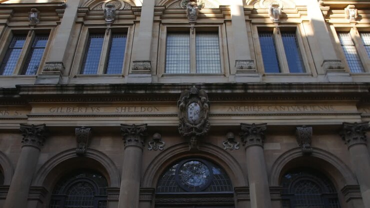 Facciata di un edificio all'Università di Oxford con colonne classiche e figure scolpite sopra alte finestre, catturata dal basso con la luce del sole che proietta ombre sulla muratura dettagliata.