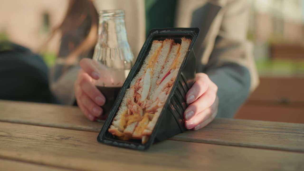 Close up of sandwich pack filled with meat and cheese placed on wooden table beside glass bottle of juice while hands move it, warm outdoor setting with natural daylight atmosphere
