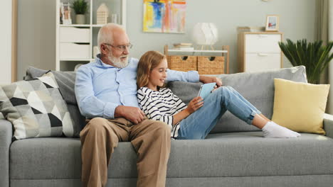 Pequeña-Adolescente-Con-El-Pelo-Largo-Y-Rubio-Descansando-En-El-Sofá-Con-Su-Abuelo-Con-Gafas-Y-Mostrándole-Algo-En-La-Pantalla-De-La-Tableta-En-La-Sala-De-Estar