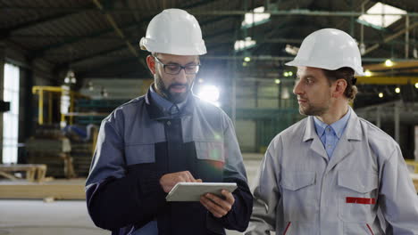 two engineers wearing helmets holding tablet while talking and walking in a factory