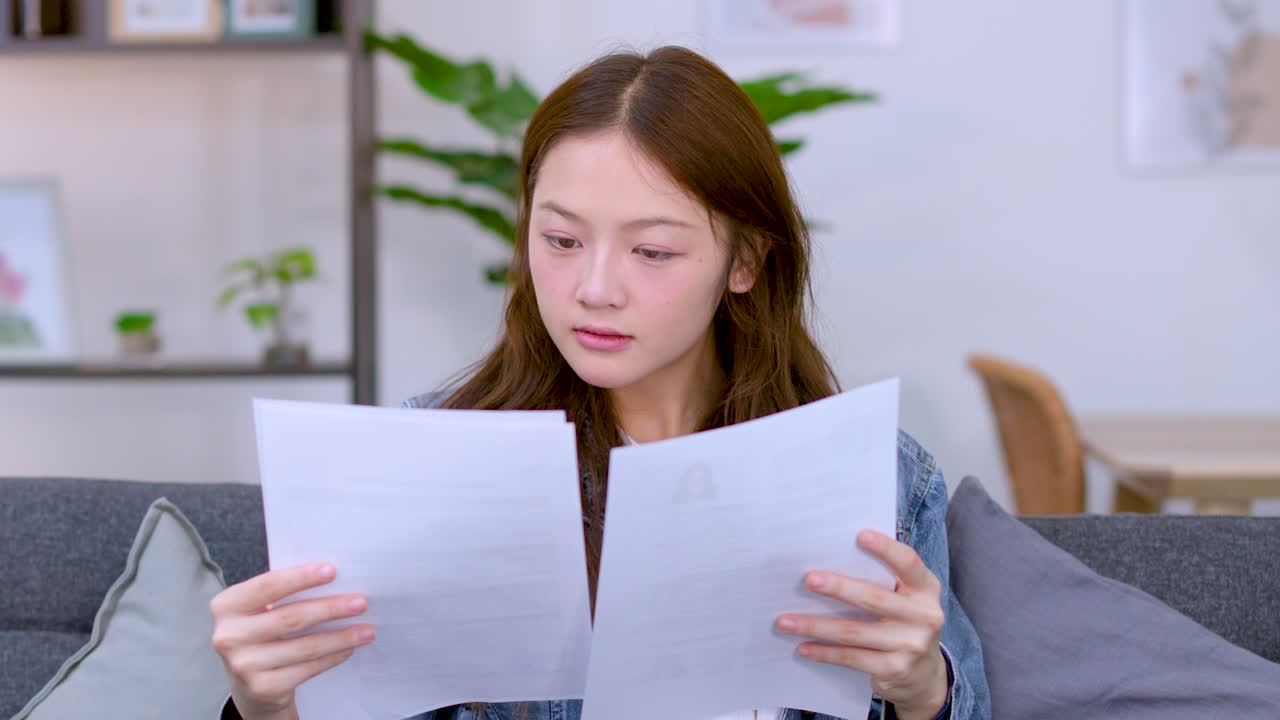 若いアジア人女性がソファに座り、幸せそうで集中した表情で書類に目を通しています。