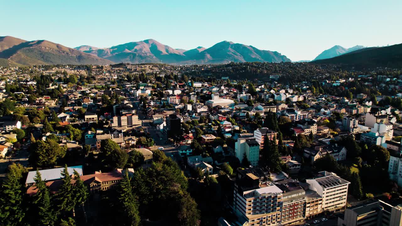 Wide-angle drone shot capturing the dense residential areas and urban development of San Carlos de Bariloche, Argentina, contrasting with the vast, rugged slopes of the Patagonian Andes mountains