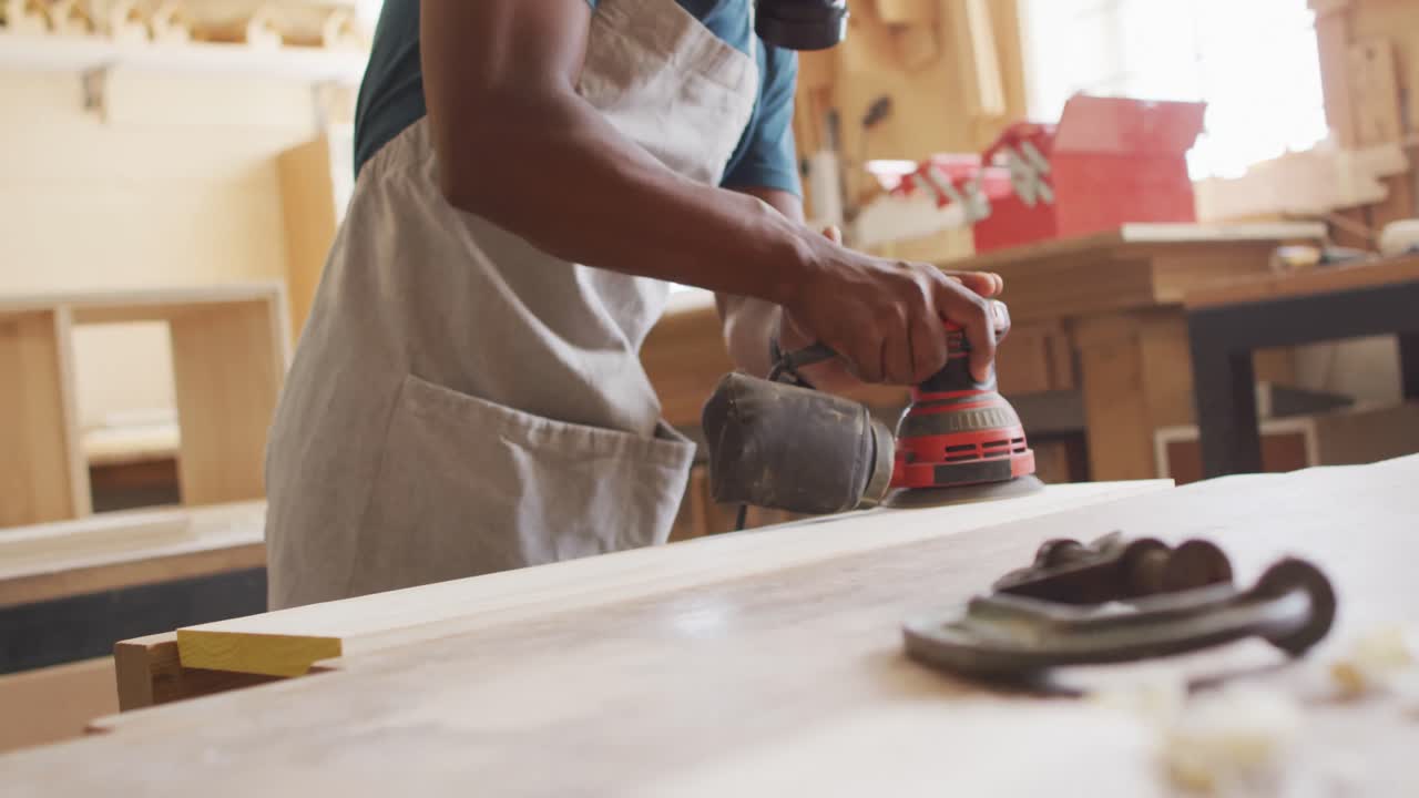 아프리카계 미국인 남성 목수 는 보호 마스크 를 착용 하여 나무 판 을 전기 산더 로 산화 하고 있다