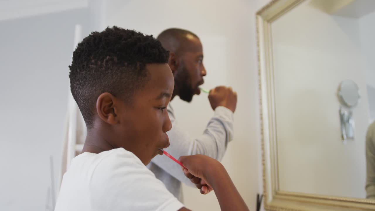 African american dad and son brushing teeth together looking in the mirror at home