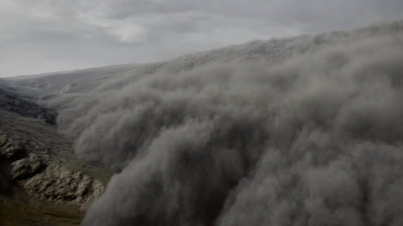Densa nube de polvo fluyendo y cayendo por una ladera escarpada después de cruzar la cresta, siguiendo la pendiente.