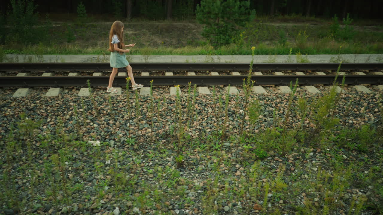 Side view of little girl walking beside railway sleeper holding grass strand in hand, wearing denim dress and striped shirt, surrounded by gravel stones and wild plants