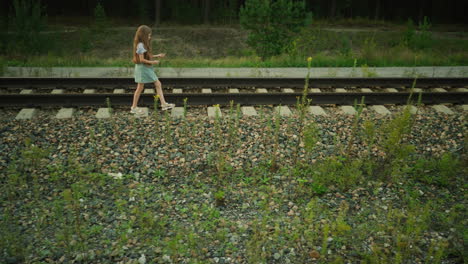side view of little girl walking beside railway sleeper holding grass strand in hand, wearing denim dress and striped shirt, surrounded by gravel stones and wild plants