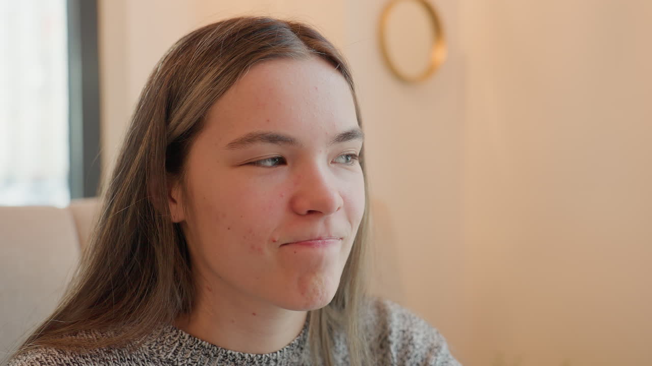 Young woman in cafe blinking eyes, smiling with mouth folded, and turning head sideways in a playful, cheerful expression while seated, radiating warmth and friendliness in casual attire