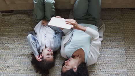 Happy-mixed-race-mother-and-daughter-laying-on-the-floor,having-fun-and-using-tablet