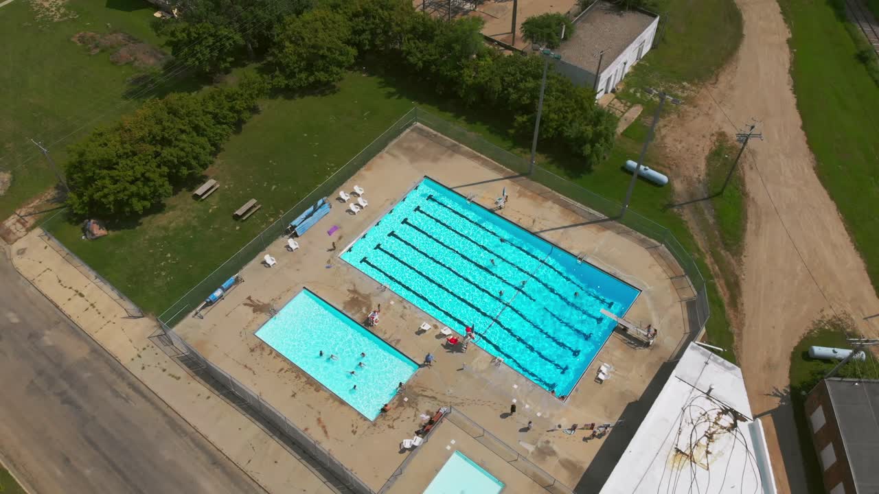 Ampla foto aérea em órbita e ângulo descendente de um complexo de piscinas públicas ao ar livre em uma cidade pequena, com uma piscina principal e piscinas menores adjacentes visíveis abaixo enquanto os nadadores se movem pelas raias