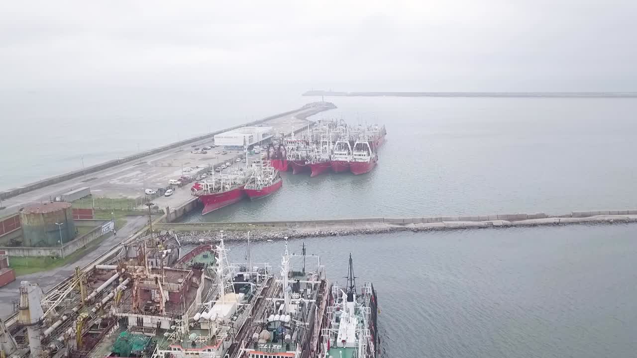 Drone flying over several fishing ships docked at an Argentine port on a cloudy day.