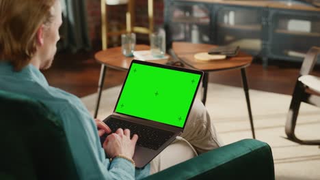 joven adulto trabajando desde su apartamento de loft en una computadora portátil con pantalla verde. hombre creativo navegando por la web, escribiendo en las redes sociales. imágenes cercanas sobre el hombro.