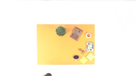 Top-down-view-of-workspace-with-a-notebook,-calendar,-paper,-envelope,-pencil-case,-stamp,-small-plant,-copy-space-on-yellow-paper-on-white-background-with-woman’s-hand-placing-the-items