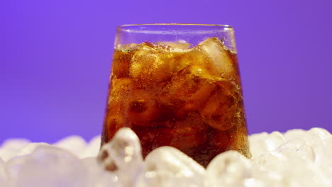 close up de una persona vertiendo una bebida fría en un vaso en cubos de hielo contra un fondo púrpura