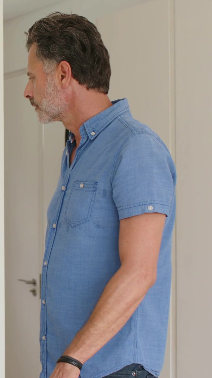Video verticale: Un uomo maturo con una camicia blu e un orologio entra in una porta e abbassa una scatola per appoggiarla.