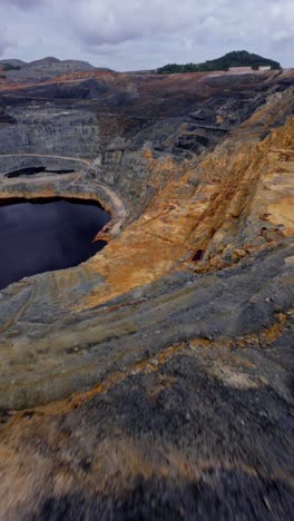 drone flying over colorful terraced rock walls of an open-pit mine excavation, dark tailings pond, barrick gold, cotuí, mining industry, environment, dominican republic. aerial fpv, vertical format