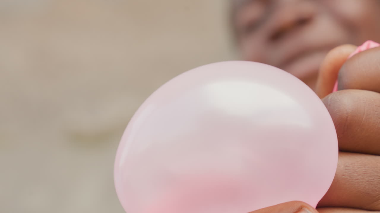 Ragazzo di colore che gonfia un palloncino rosa, primo piano di bocca e guance, sforzo visibile e respiro concentrato mentre il bambino gonfia il giocattolo all'aperto contro un muro di cemento, energia spontanea e determinazione giocosa.