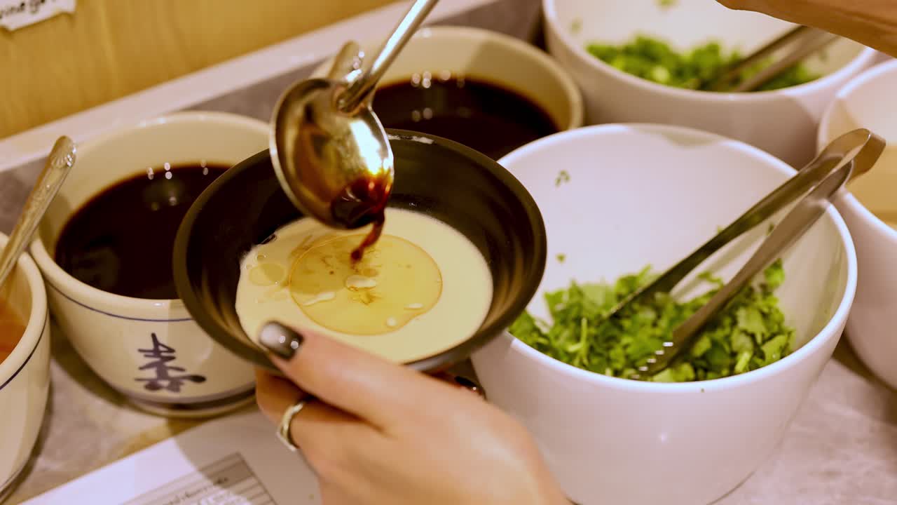 Hand ladles chili oil into sesame sauce bowl at bright, organized Chinese hotpot condiment station