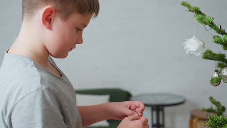 Close view of boy in grey top standing in front of christmas tree holding ornament in hand, preparing to decorate, festive indoor setting, joyful holiday atmosphere, bright and cozy background