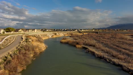 Imágenes-Aéreas-De-Un-Río-Que-Fluye-Hacia-Las-Montañas-Distantes-Y-Las-Nubes-A-Lo-Largo-De-Un-Sendero-Para-Bicicletas-Pavimentado