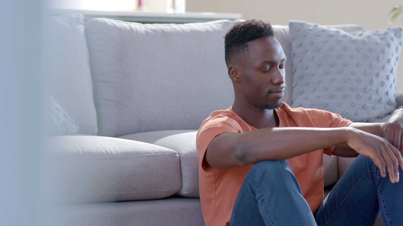 Worried african american man sitting and thinking in sunny living room, slow motion