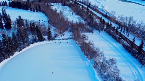 4k winter aerial flyover drop sunset reflects shadow trees snow covered skate path next to roadside access parked cars connected to largest outdoor manmade ice rink adults skating edm victoria park5-6