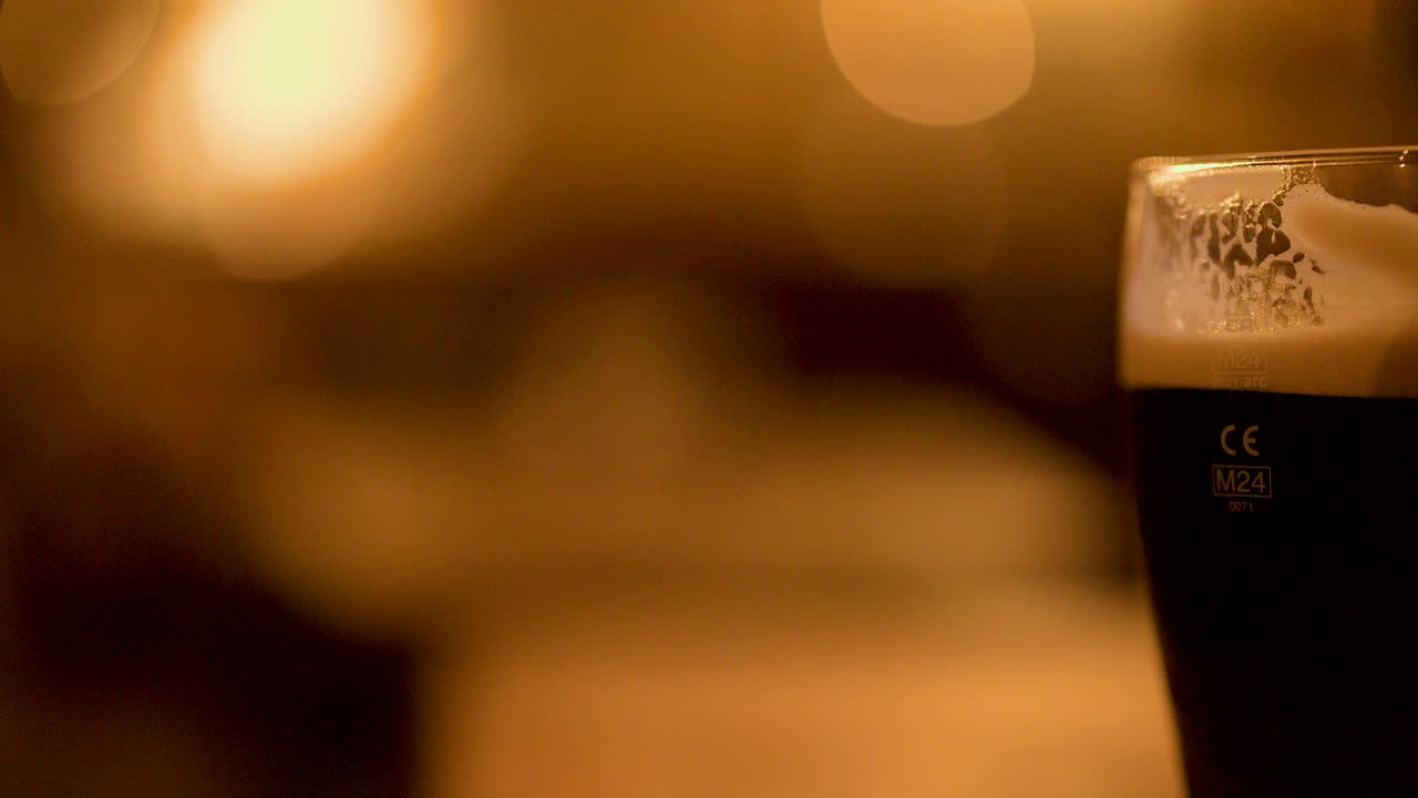 Hand raises dark beer pint glass in warmly lit pub, shallow focus, evening ambiance
