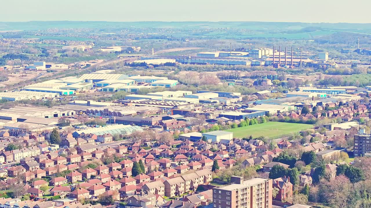 Panoramic aerial showing dense residential terraces in foreground and a large industrial estate with warehouses, factory buildings, and multiple chimneys or smokestacks beyond, hills on the horizon
