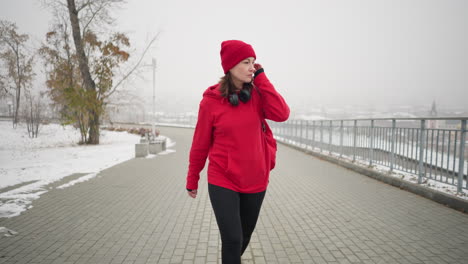 athlete wearing red jacket walking along interlocked pathway with bag over shoulder, gazing away thoughtfully, near iron railing and blurred cityscape view in foggy winter atmosphere