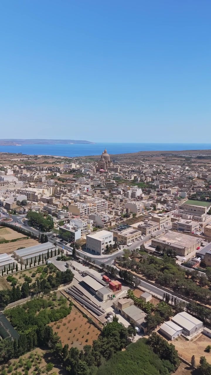Rotonda di Xewkija e Chiesa di San Giovanni Battista a Gozo, Malta, circondate da edifici residenziali, strade e paesaggio mediterraneo sotto un cielo blu in una veduta aerea della città
