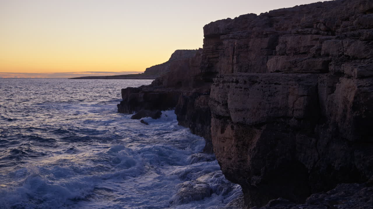 Dramatic coastal cliffs at Cape Greco, Cyprus, during sunset. Waves crash against rugged limestone rocks under a warm golden sky