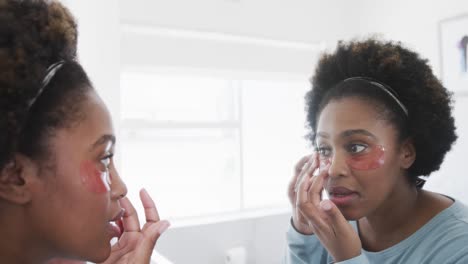 happy african american woman applying under eye masks looking in bathroom mirror, slow motion