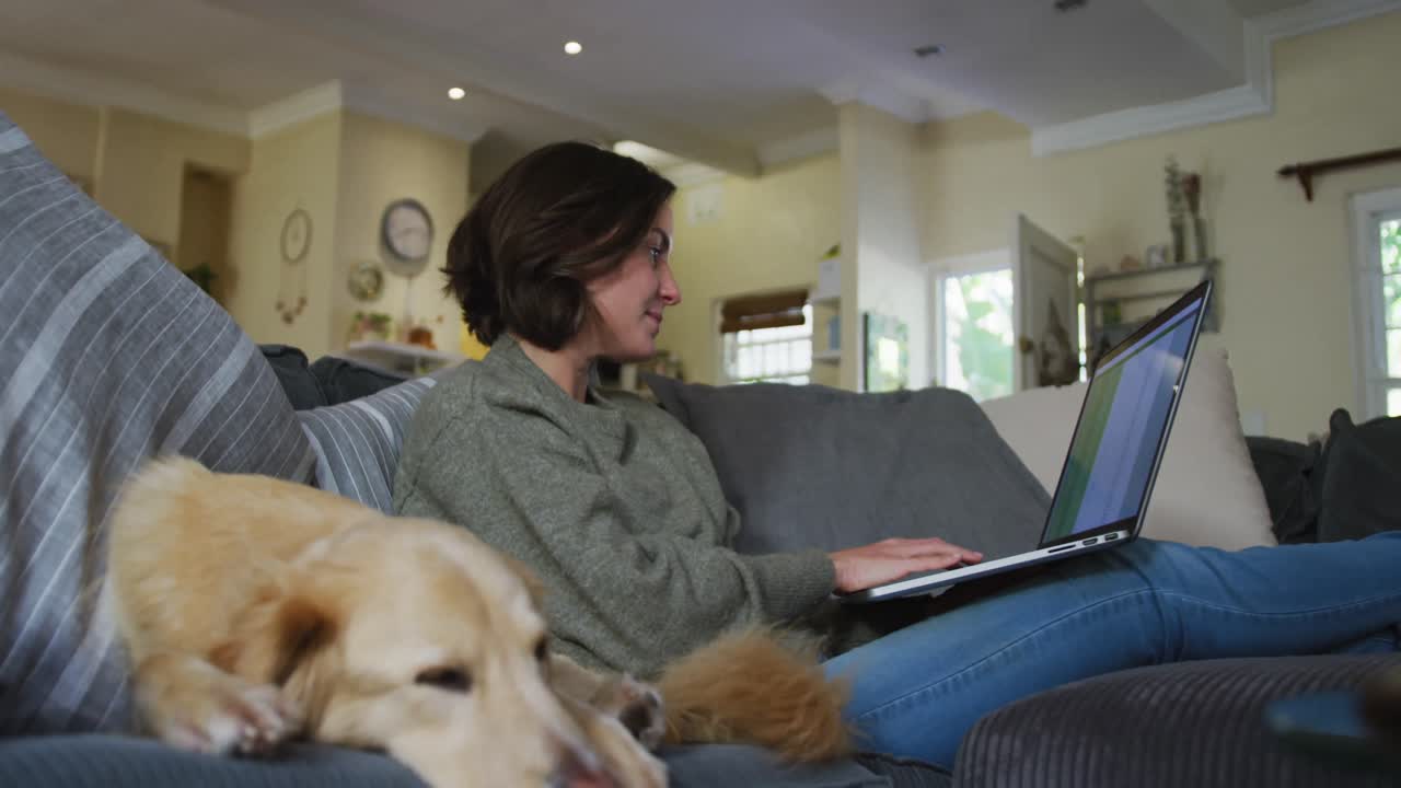 Smiling caucasian woman working from home and stroking her pet dog on sofa next to her