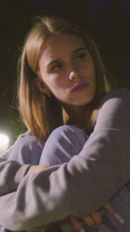 dama sentada en un banco del parque por la noche con los brazos cruzados sobre las rodillas, descansando la cabeza pensativamente, mirando a la distancia bajo la suave iluminación del parque, expresando calma contemplación y estado de ánimo reflexivo