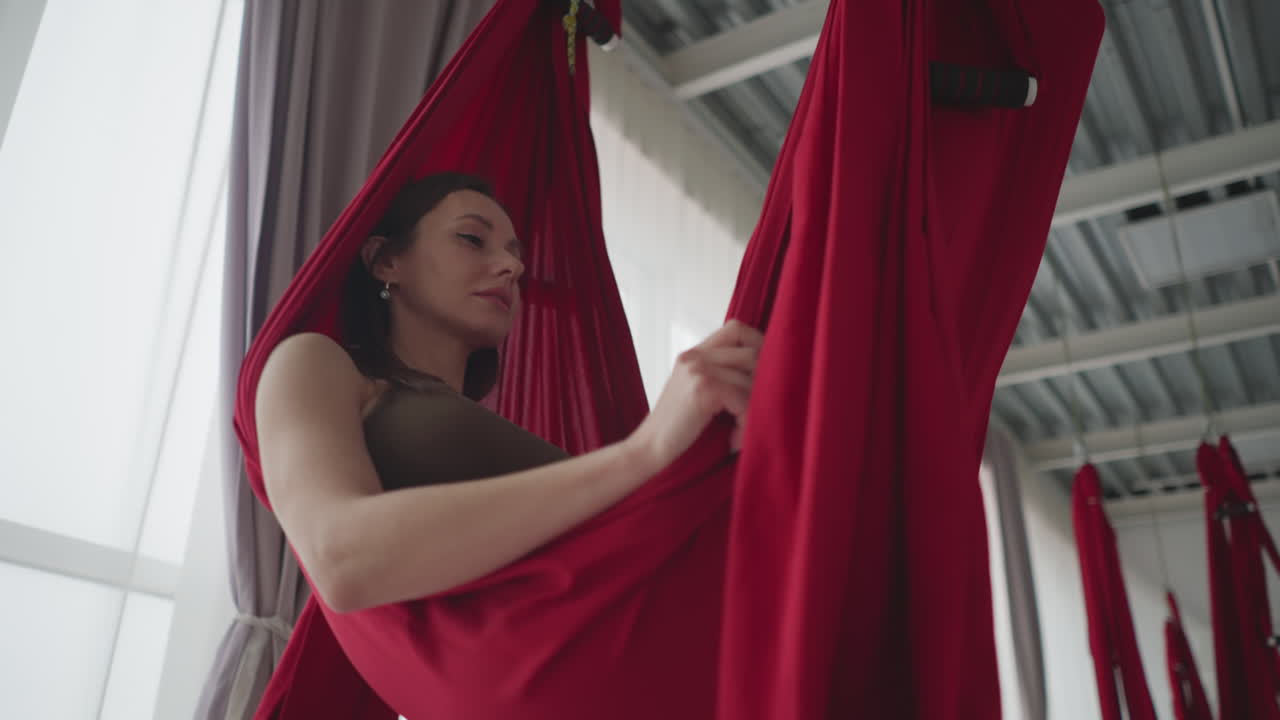 Low angle view of wellness guide lying peacefully in red silk hammock swinging gently in bright yoga studio with sheer curtains and urban view conveying calm balance focus and serene flow practice