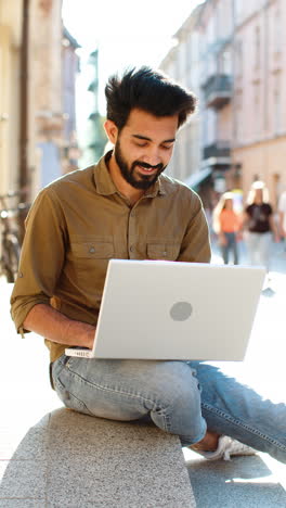 indian man freelancer working online distant job with laptop sitting in city street browsing website
